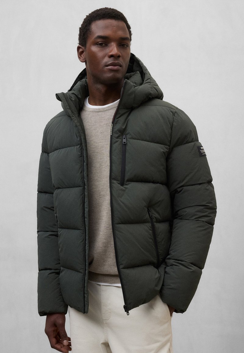 Mørkegrøn dunjakke med hætte, med en lynlåslomme på brystet, quiltet design og tekstureret stof. Båret over en lyse trøje.