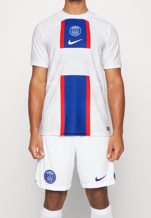 Mężczyzna ubrany w biały strój piłkarski Paris Saint-Germain z niebieskimi i czerwonymi pionowymi paskami oraz logo Nike na koszulce i spodenkach.