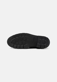 Semelle de chaussure noire avec des crampons profonds et des motifs de grip circulaires conçus pour la traction et la durabilité, présentée sur un fond uni.