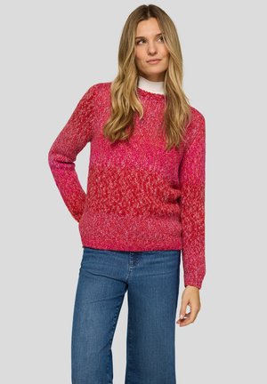 Frau mit langen blonden Haaren, die einen roten und pinken Strickpullover über einem weißen Oberteil trägt, kombiniert mit blauen Jeans, stehend vor einem grauen Hintergrund.