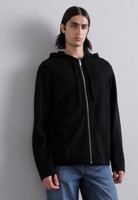 Schwarze, mit Reißverschluss versehene Kapuzenjacke mit weicher Textur, ausgestattet mit einer Kapuze, Kordeln und langen Ärmeln. Kombiniert mit blauen Jeans.