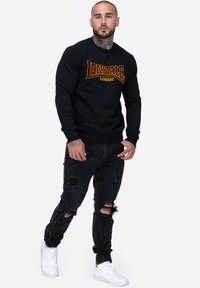 Sudadera negra con el logo en rojo y amarillo de "Lonsdale London", combinada con jeans negros desgastados y zapatillas blancas. Fondo liso.