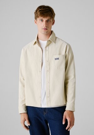 Crème corduroy shirt met een wijde kraag, knoopsluiting en borstzak met "Red Bull Racing" geborduurd in marineblauw. Draagt over een wit shirt.
