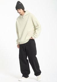 Licht beige hoodie met capuchon en trekkoord, voorzak, gecombineerd met zwarte wijde broek en crèmekleurige sneakers. Textured grijze gebreide beanie met logo.