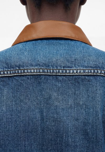 Πίσω όψη ατόμου που φοράει μπλε τζιν μπουφάν με καφέ δερμάτινο γιακά σε λευκό φόντο.