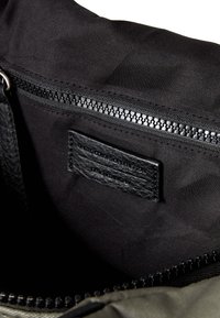 Interiér černé textilní tašky s uzávěrem na zip, texturovanou koženou etiketou a hladkým syntetickým podšívkou. Barevné kontrasty mezi materiály.