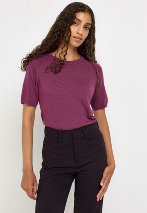 Donna con capelli lunghi e ricci che indossa un maglione viola a maniche corte e pantaloni neri a vita alta, in piedi su uno sfondo bianco.