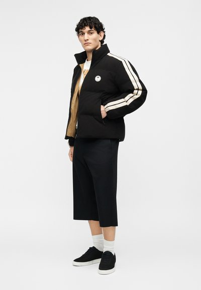 Νέος άνδρας που στέκεται φορώντας μαύρο πουπουλένιο μπουφάν με λευκές ρίγες, μαύρο σορτς με φαρδιά πόδια, λευκές κάλτσες και μαύρα αθλητικά παπούτσια.