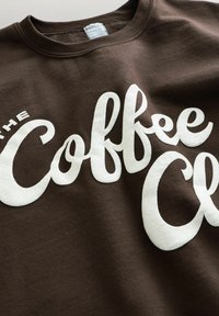 Barna pulóver fehér, vastag betűs írott felirattal: "THE Coffee." A szövet puha és sima tapintású, klasszikus kerek nyakkivágással.