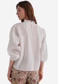 Blusa bianca di lino con maniche a sbuffo, bordo in pizzo sullo scollo e retro con apertura a forma di chiave. Abbinata a pantaloni ricamati floreali e a fantasia.