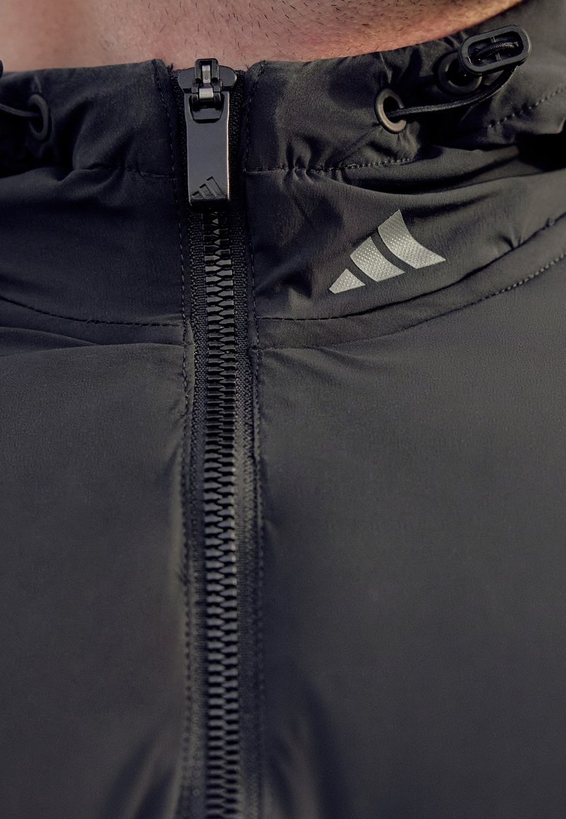 Nahaufnahme einer schwarzen Zip-Jacke mit Zugbändern und einem reflektierenden Adidas-Logo im Brustbereich.