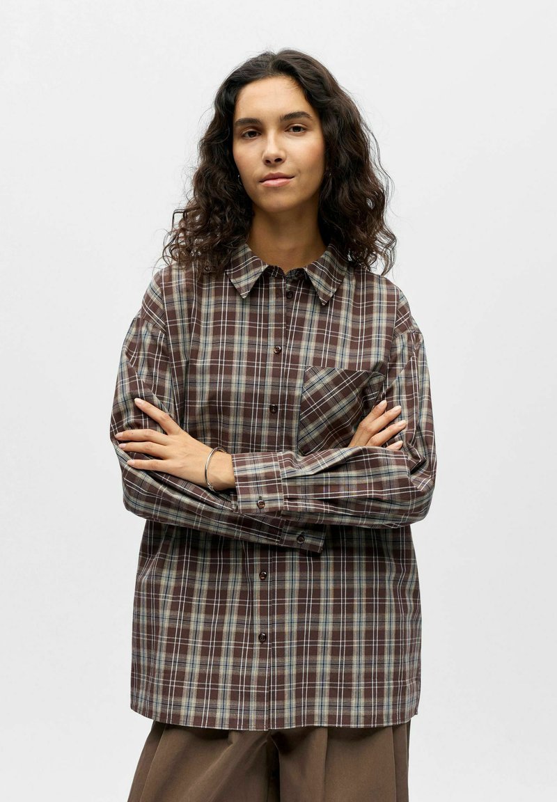 Chemise à carreaux marron avec col boutonné, poche avant et coupe ample. Le tissu présente une texture lisse avec un mélange de tons bleus et beiges.