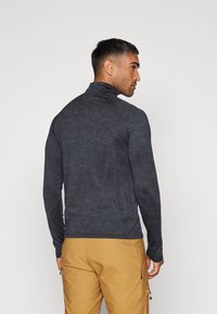Top de manga larga gris oscuro con una textura sutil, cuello alto y un diseño ajustado, combinado con pantalones beige que tienen bolsillos con cremallera.