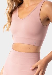 Sujeta de deporte de color rosa claro, hecha de un tejido suave y elástico, con un diseño de escote en V, banda acanalada y un sutil detalle de logo en la parte inferior.
