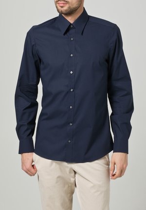 Homme portant une chemise cintrée bleu foncé à manches longues avec col pointu et un pantalon beige, debout devant un fond uni.