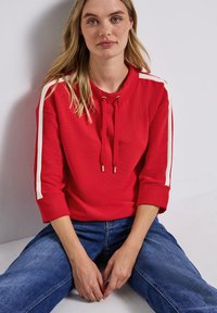 Pullover rosso con colletto rotondo e coulisse, caratterizzato da stripe laterali bianche. Tessuto a coste, maniche a tre quarti e vestibilità rilassata.
