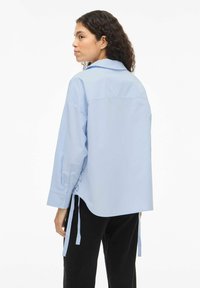 Chemise surdimensionnée bleu clair avec un empiècement dans le dos, manches longues et liens latéraux. Confectionnée en tissu lisse avec des poignets à boutons.