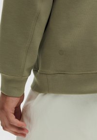 Sweatshirt vert olive avec une coupe décontractée, tissu texturé, poignets côtelés et un petit logo en bas. Main visible sur un bas de couleur claire.