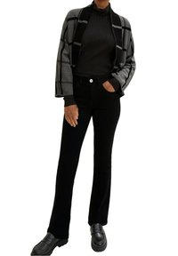 Schwarze Hose mit weitem Bein, kombiniert mit einer grau-schwarz karierten Strickjacke mit Rippenstruktur und einem eng anliegenden Rollkragenpullover darunter. Schwarze Loafers.