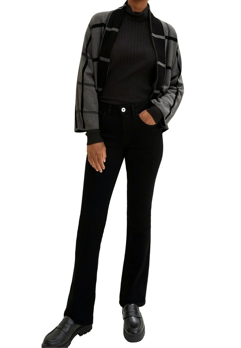 Schwarze Hose mit weitem Bein, kombiniert mit einer grau-schwarz karierten Strickjacke mit Rippenstruktur und einem eng anliegenden Rollkragenpullover darunter. Schwarze Loafers.