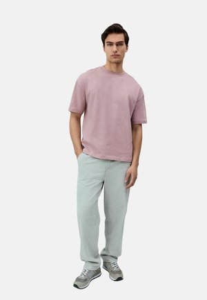 Camisa de manga corta rosa claro, corte relajado, combinada con jeans verde claro. El modelo lleva zapatillas grises con acentos negros. Diseño simple y moderno.