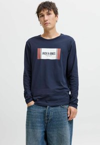 Fiatal férfi sötétkék, hosszú ujjú pólóban a "JACK & JONES" logójával és széles kék farmernadrágban, kezével a zsebében áll egy sima háttér előtt.
