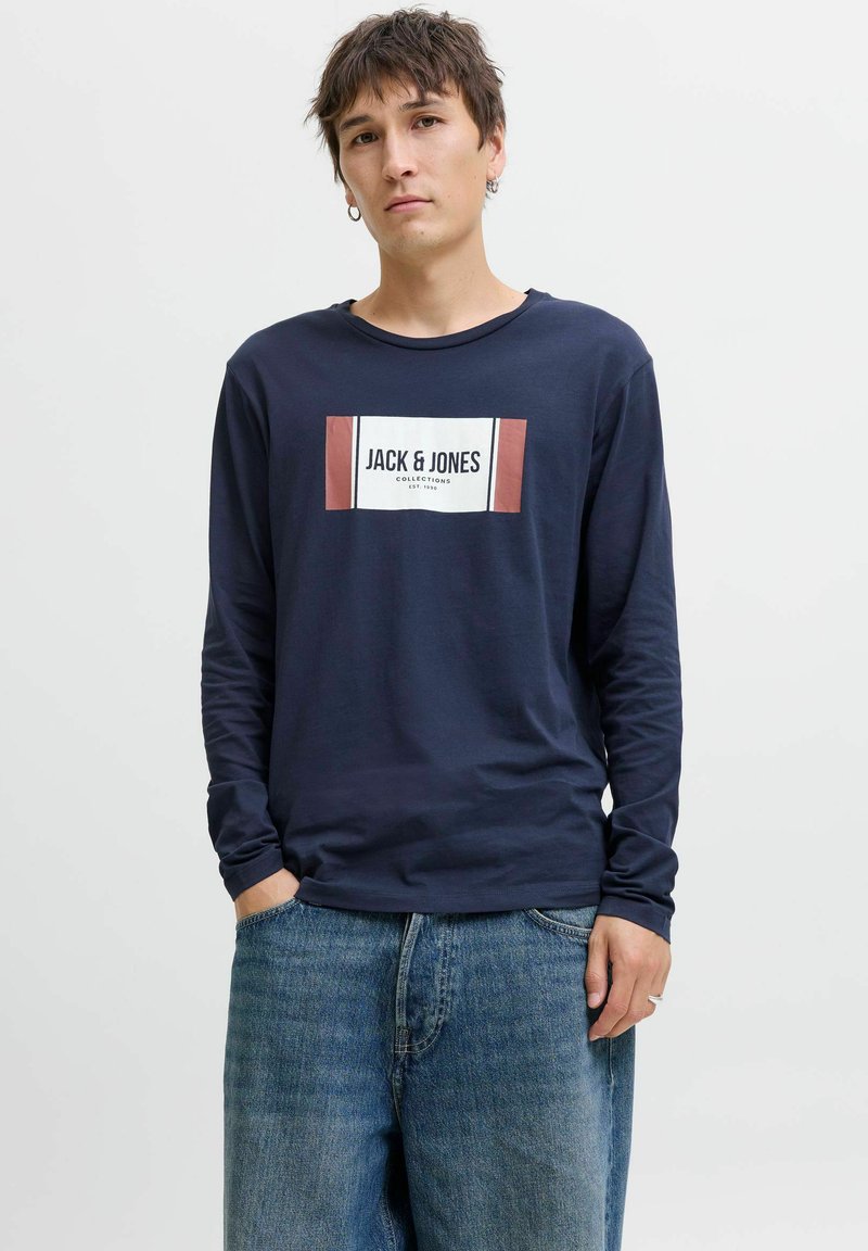 Fiatal férfi sötétkék, hosszú ujjú pólóban a "JACK & JONES" logójával és széles kék farmernadrágban, kezével a zsebében áll egy sima háttér előtt.