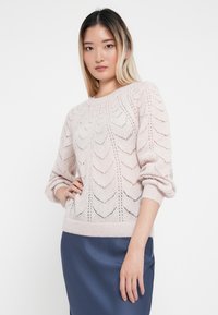 Jasnoróżowy sweter dziany z wzorzystym otwartym splotem, okrągłym dekoltem i długimi, lekko bufiastymi rękawami, noszony na ciemnoniebieskiej spódnicy.