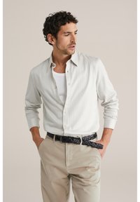 Leicht gestreiftes Langarmhemd in Grau, aus weichem Stoff. Über einem weißen Tanktop getragen und kombiniert mit beigen Hosen sowie einem schwarzen geflochtenen Gürtel.