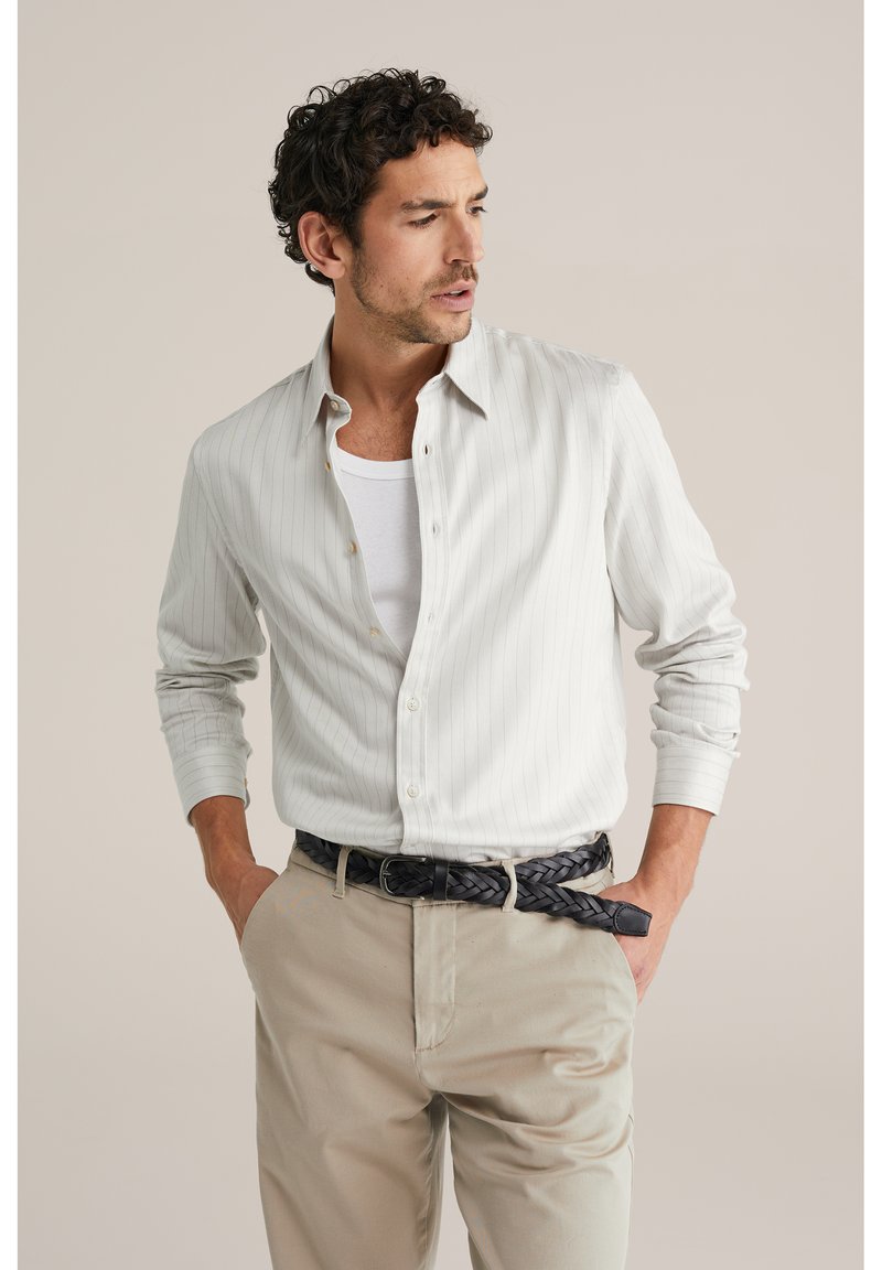 Leicht gestreiftes Langarmhemd in Grau, aus weichem Stoff. Über einem weißen Tanktop getragen und kombiniert mit beigen Hosen sowie einem schwarzen geflochtenen Gürtel.