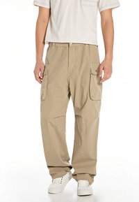 Pantalones cargo beige con múltiples bolsillos laterales, cinturilla elástica y corte relajado. Llevados con zapatillas blancas y una camisa de color claro.