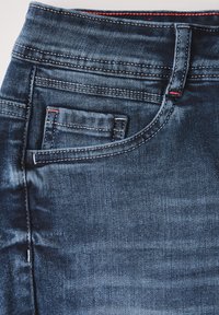 Shorts en denim bleu foncé avec une texture lisse, des coutures visibles et une petite poche avant avec des surpiqûres rouges.
