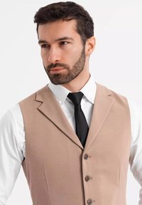 Hellbeige Weste aus strukturiertem Stoff, mit einem Revers, drei Knöpfen und einer schwarzen Krawatte über einem weißen Hemd mit Kragen.