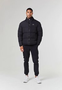 Crna puhasta jakna s horizontalnim prošivenim dizajnom, visokim ovratnikom i patentnim zatvaračem, uparena s crnim cargo hlačama i bijelim tenisicama.