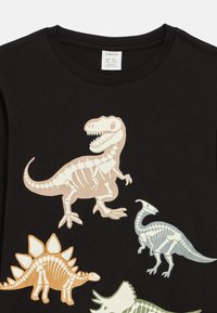 Cămașă neagră cu patru ilustrații colorate ale scheletelor de dinozauri: T-Rex, Stegosaurus, Parasaurolophus și Triceratops.