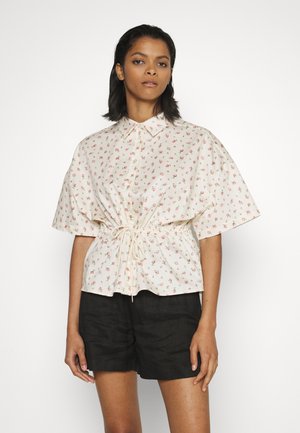 Blusa floral clara com mangas curtas, cintura franzida com cordão, frente com botões e fundo creme. Combinada com calções escuros.