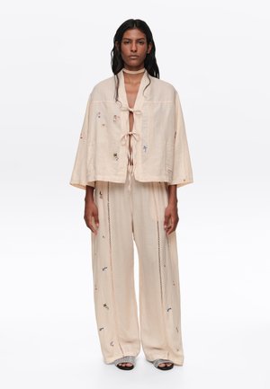 Mujer con conjunto suelto beige con pequeños detalles bordados y coloridos, parte superior con apertura frontal atada con dos lazos, pantalones de pierna ancha y zapatos abiertos en los dedos.