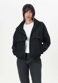 Μαύρο ελαφρύ μπουφάν με μπροστινό φερμουάρ, με κουμπιά και δύο μπροστινές τσέπες. Διαθέτει ψηλό γιακά και ελαστικές μανσέτες, συνδυασμένο με ένα λευκό πουκάμισο.