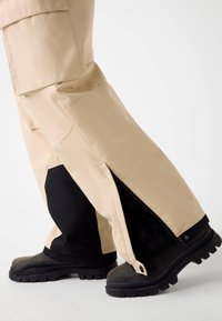 Beige Cargo-Hosen mit schwarzen Akzenten, ausgestattet mit seitlichen Reißverschlüssen und einer lockeren Passform, kombiniert mit robusten schwarzen Stiefeln mit einem groben Profil.