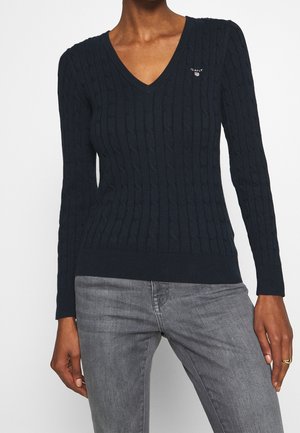 Personne portant un pull bleu marine en maille torsadée avec col en V et un jean gris, avec un petit logo de marque sur la poitrine et une bague en or à la main droite.