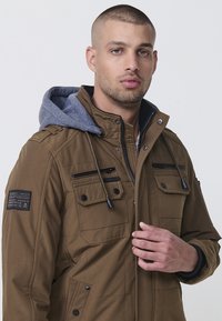 Braune gepolsterte Jacke mit einer grauen Kapuze. Verfügt über mehrere Reißverschlusstaschen, einen hohen Kragen und Kordelzugdetails. Glatte Stoffoberfläche.