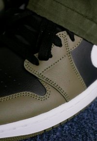 Black and olive green sneaker with a smooth leather finish, round toe, black laces, and white logo. Features visible stitching and rubber sole.