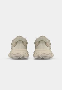 Ett par beige sportskor sett bakifrån med texturerad sula och dragflikar mot en vit bakgrund.