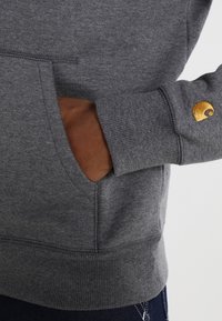 Mano nella tasca di una felpa grigia con logo dorato ricamato sul polsino della manica, indossata con jeans blu scuro.