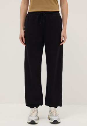 Persona che indossa pantaloni della tuta neri con polsini elastici, top beige e scarpe da ginnastica bianche, in piedi davanti a uno sfondo chiaro e uniforme.