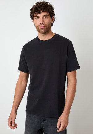 Homme aux cheveux bouclés portant un t-shirt noir uni à manches courtes et un jean foncé, debout devant un fond clair uni.