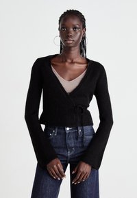 Svart omslagstrøye med lange, utsvingte ermer, laget av myk stoff. Stylet over en beige tettsittende topp, paret med mørkeblå denimjeans.