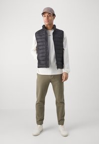 Svart quiltad puffer-väst med hög krage, över en ljusfärgad långärmad skjorta. Bärs med olivgröna byxor och vita sneakers.