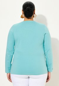 Türkise Langarm-T-Shirt aus weichem Stoff, mit rundem Ausschnitt und lockerer Passform, ergänzt durch weiße Hosen.