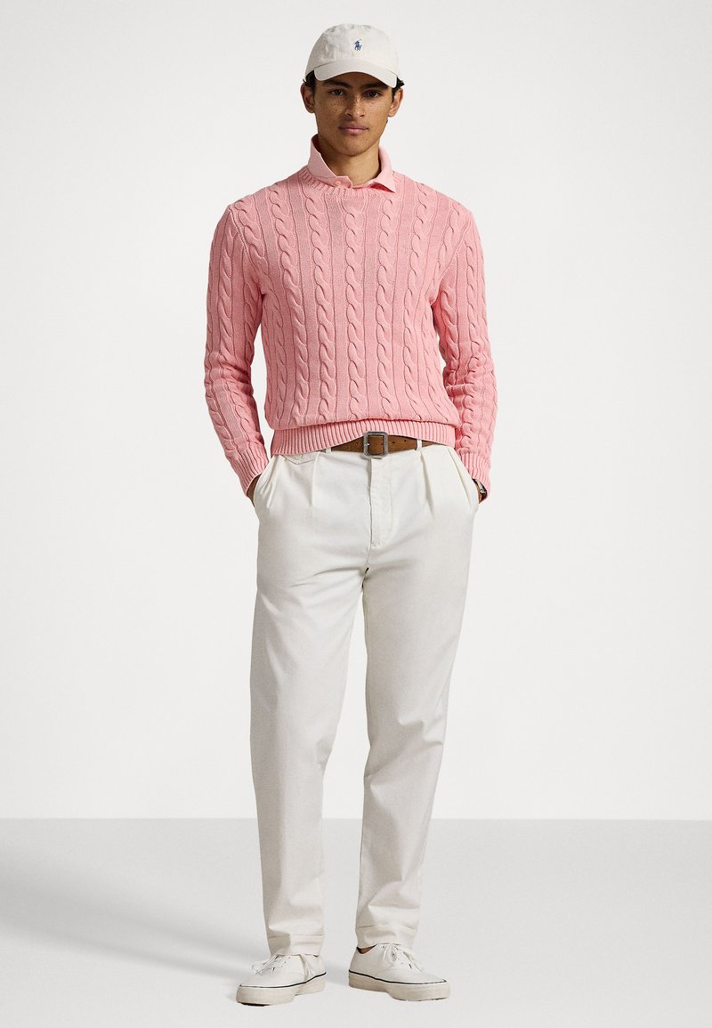 Jasnoróżowy sweter w splot kablowy, kremowe spodnie i białe sneakersy. Modelka ma na sobie białą czapkę z niebieskim logo i prezentuje swobodny styl.